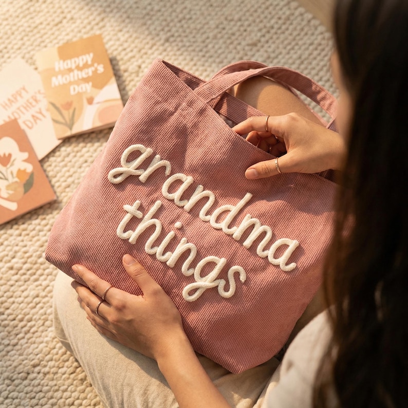 May include: A dusty rose corduroy tote bag featuring the white embroidered text "grandma things" in a cursive font. The bag has two fabric handles and a textured ribbed surface, showcasing a soft pink color palette and a casual design.