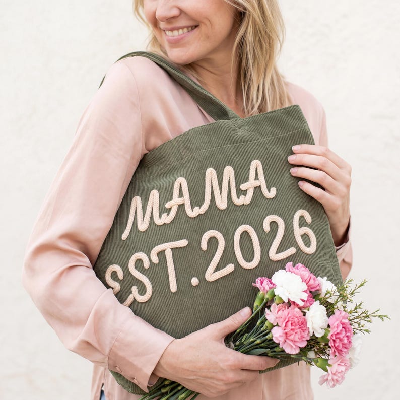May include: An olive green corduroy tote bag featuring the cream-colored embroidered text MAMA EST. 2026 on the front. The bag has a single shoulder strap and is held alongside a small bouquet of pink and white carnations with green foliage.