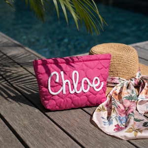 May include: A vibrant pink quilted cosmetic bag, patterned with hearts, featuring the name "Chloe" in white rope lettering. Beside it are a straw hat and a floral scarf, all on a wooden surface.