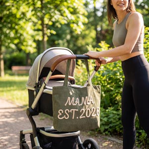 May include: An olive green corduroy tote bag hanging from the handle of a gray and black baby stroller. The bag features the white embroidered text MAMA EST. 2026 in a cursive font. The stroller has a brown leather-look handle and a storage basket.