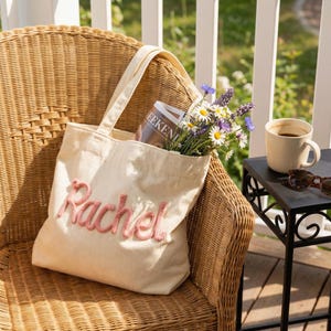 May include: A cream-colored corduroy tote bag featuring the name Rachel embroidered in raised pink yarn lettering. The bag contains a magazine and a bouquet of wildflowers including daisies, lavender, and blue cornflowers, with two shoulder straps.