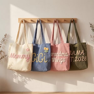 May include: Four canvas tote bags hanging on a wooden rack. The cream bag says Mommy with a flower, the blue bag says GiGi with a flower, the pink bag says Grandma things, and the olive green bag says MAMA EST. 2026 in raised fabric lettering.