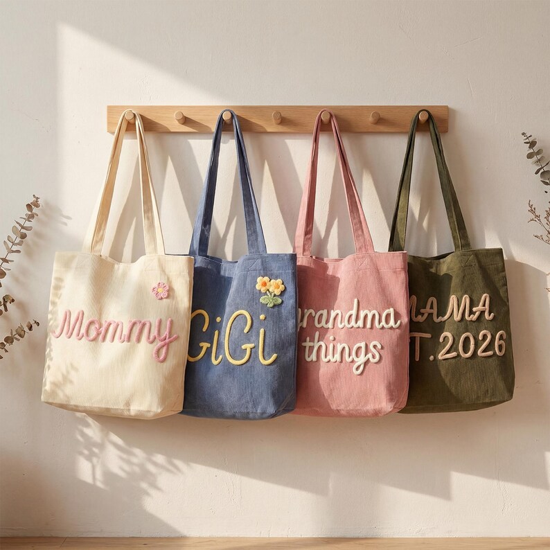 May include: Four canvas tote bags hanging on a wooden rack. The cream bag says Mommy with a flower, the blue bag says GiGi with a flower, the pink bag says Grandma things, and the olive green bag says MAMA EST. 2026 in raised fabric lettering.