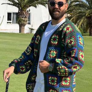 Peut inclure: Un cardigan crocheté vert, bleu, rouge et jaune. Le pull sans fermeture présente un motif de carrés granny. La personne porte un t-shirt blanc et un pantalon blanc. L'arrière-plan comprend des palmiers et un terrain de golf.