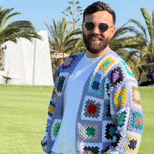 Peut inclure: Un cardigan en crochet multicolore avec un motif patchwork. Le pull sans fermeture présente des carrés de différentes couleurs, dont le bleu, le jaune, le rouge et le vert, porté sur un t-shirt blanc. L'arrière-plan montre une pelouse verte et des palmiers.
