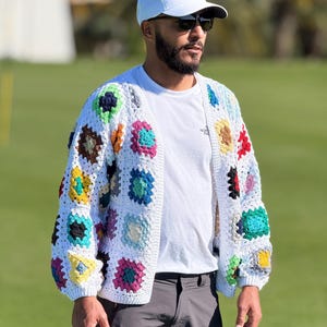 Peut inclure: Un cardigan blanc en crochet avec des motifs carrés colorés. Le pull sans fermeture présente une variété de couleurs vives, dont le rose, le bleu, le jaune et le vert. La personne porte une casquette blanche et des lunettes de soleil.