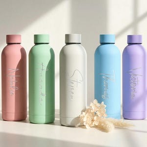 Peut inclure: Cinq gourdes colorées, rose, vert, blanc, bleu et violet. Chaque gourde a un bouchon assorti et un nom en script argenté. Un petit bouquet de fleurs séchées est devant.