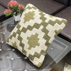 Könnte beinhalten: Quadratisches Zierkissen mit einem geometrischen Muster in Olivgrün und Creme. Das Kissen besteht aus einem strukturierten Material. Es liegt auf einem Glastisch. Eine kleine Topfpflanze mit rosa Blüten befindet sich im Hintergrund.
