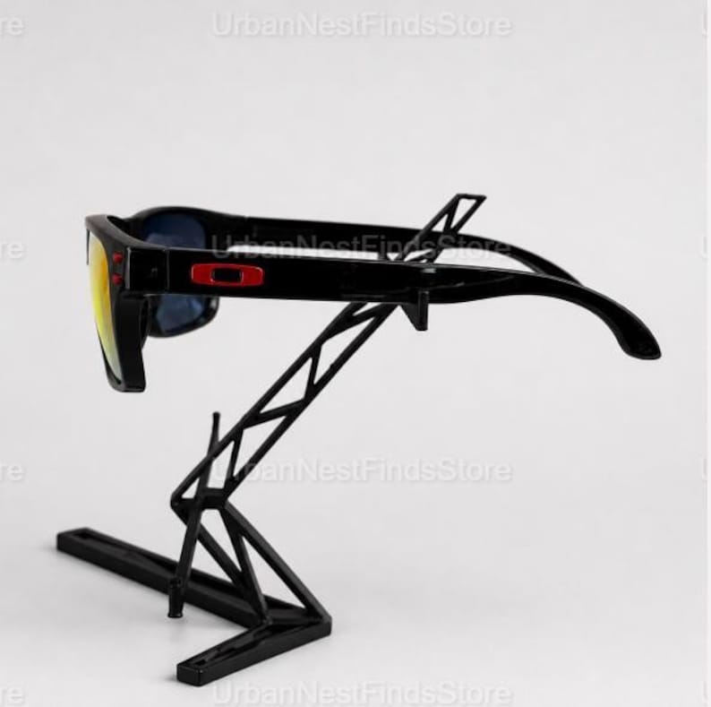Puede incluir: Gafas de sol negras con lentes amarillas y azules se exhiben en un soporte de metal negro. Las gafas de sol tienen un logotipo rojo en el lateral. El soporte tiene un dise&ntilde;o geom&eacute;trico.