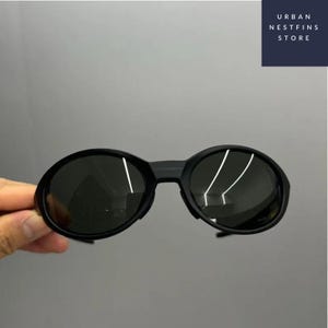 Puede incluir: Gafas de sol ovaladas negras con lentes oscuras. Las gafas tienen una montura gruesa y un puente que conecta las dos lentes. El texto "URBAN NESTFINS STORE" está en un rectángulo azul marino en la esquina superior derecha.