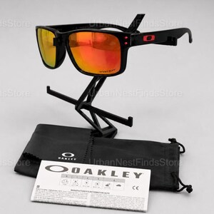 Puede incluir: Gafas de sol Oakley negras con lentes naranjas, expuestas sobre un soporte negro. Las gafas tienen una montura cuadrada y el logotipo de Oakley en la patilla. Una funda negra y una nota informativa del fabricante est&aacute;n debajo.