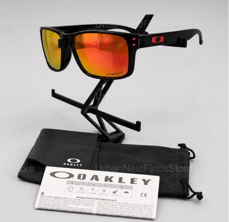 Puede incluir: Gafas de sol Oakley negras con lentes naranjas, expuestas sobre un soporte negro. Las gafas tienen una montura cuadrada y el logotipo de Oakley en la patilla. Una funda negra y una nota informativa del fabricante est&aacute;n debajo.