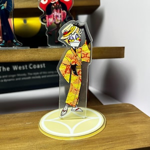 May include: A clear acrylic figure of a cartoon character in a yellow kimono with red and orange accents, a straw hat, and black gloves. The figure stands on a yellow circular base with a decorative design. Other figures are visible in the background.