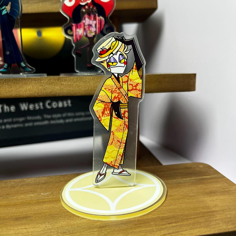 May include: A clear acrylic figure of a cartoon character in a yellow kimono with red and orange accents, a straw hat, and black gloves. The figure stands on a yellow circular base with a decorative design. Other figures are visible in the background.