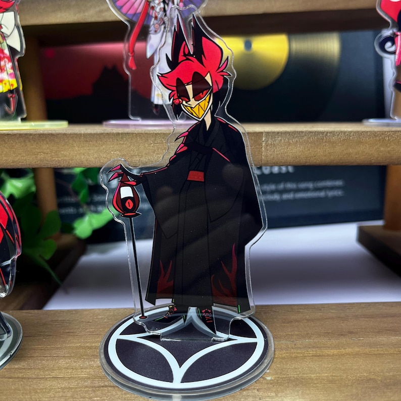 May include: A clear acrylic figure of a character with red hair, a yellow-toothed grin, and a black outfit. The figure is holding a red lantern and stands on a black and white circular base. The character has a red and black color scheme.