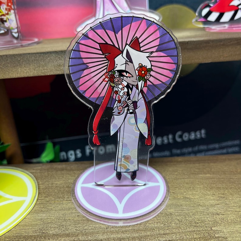 May include: A clear acrylic stand-up figure featuring a character in a kimono holding a parasol. The figure has red and white hair and is adorned with floral accents. The parasol is pink and purple. The base is a light purple circle with a white design.