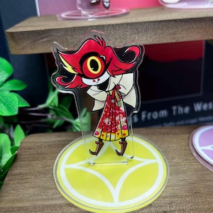 May include: A clear acrylic stand-up figure featuring a cartoon character with red hair, a large yellow eye, and a red and white patterned dress. The figure is on a yellow circular base with a white design. The figure is on a wooden shelf.