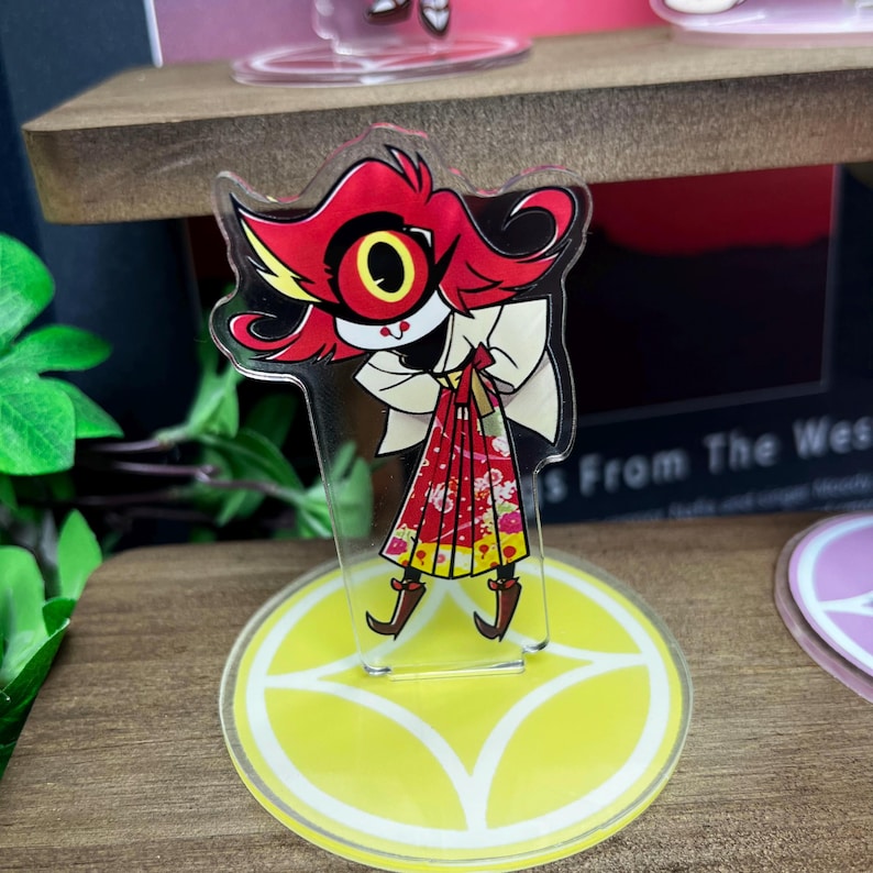May include: A clear acrylic stand-up figure featuring a cartoon character with red hair, a large yellow eye, and a red and white patterned dress. The figure is on a yellow circular base with a white design. The figure is on a wooden shelf.