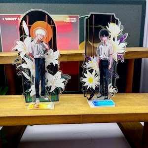 Könnte beinhalten: Zwei durchsichtige Acryl-Aufsteller mit Anime-Figuren. Die linke Figur hat weißes Haar, trägt ein weißes Hemd und eine dunkle Hose, umrahmt von weißen Lilien. Die rechte Figur hat schwarzes Haar, trägt ein weißes Hemd und eine dunkle Hose, mit weißen Blumen.