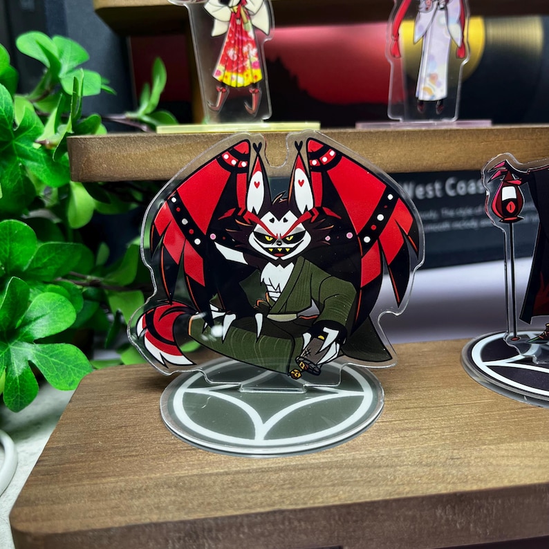 May include: A clear acrylic stand featuring a cartoon character with red and black wings, a green jacket, and a white face. The stand has a decorative base and is displayed on a wooden shelf. Other similar character stands are visible in the background.