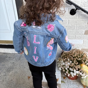 May include: A blue denim jacket featuring various colorful embroidered patches, including hearts, a milk carton, and a butterfly. The back of the jacket displays the name LIV in large, pink-bordered white letters arranged vertically down the center.