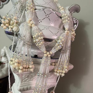 May include: A white beaded garland with cream-colored accents and sparkling crystal beads. The garland is draped over a white decorative vase with a silver top and accents. The beads are arranged in a cascading pattern.