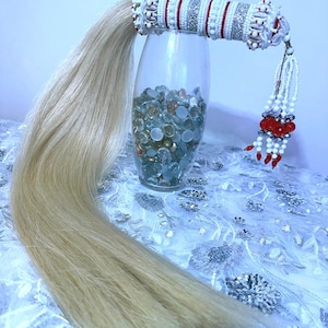 May include: A decorative hair accessory with a long, blonde ponytail. The cylindrical base is adorned with white beads, red accents, and silver embellishments. A beaded tassel with red and white beads hangs from the base. The accessory is displayed in a clear vase.