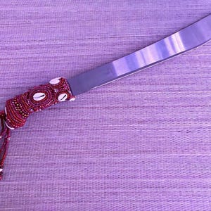 May include: A large, stainless steel knife with a decorative handle. The handle is wrapped in red and white beads and adorned with seashells. A tassel of red beads hangs from the handle. The knife rests on a purple woven mat.