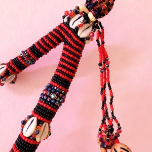 May include: A decorative staff wrapped in alternating bands of red and black seed beads, accented with white cowrie shells and iridescent beads. Two small, light-coloured, hand-painted gourds with red and black markings hang from the top by beaded cords.