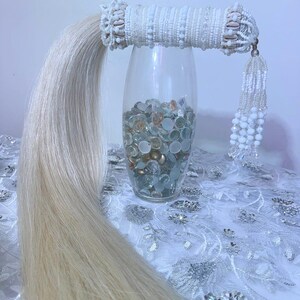 May include: A decorative hair accessory with a white beaded band and a long, flowing, light blonde hair extension. The band is adorned with small shells and a dangling beaded tassel. The hair piece is displayed next to a glass vase filled with decorative stones.