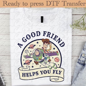 Peut inclure: T-shirt blanc avec un motif de Buzz l'Éclair et Woody de Toy Story, avec le texte "A GOOD FRIEND HELPS YOU FLY." Le design comprend des planètes et des étoiles, avec un aspect vintage et vieilli.