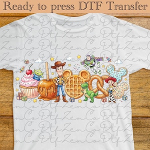 Peut inclure: T-shirt blanc avec un motif de transfert DTF coloré représentant des personnages de dessins animés, dont Woody, Buzz l'Éclair et Jessie. Le motif comprend également un cupcake, une pomme au caramel, une gaufre, un bretzel et un biscuit en forme de Mickey. Le texte en haut indique "Ready to press DTF Transfer."