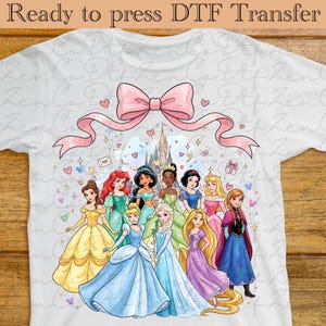 Pode incluir: Camiseta branca com um design de transferência DTF. Apresenta um laço rosa e várias princesas de desenhos animados em vestidos coloridos, com um castelo ao fundo. O texto na parte superior diz "Ready to press DTF Transfer."