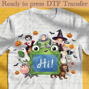 Peut inclure: Un t-shirt blanc avec un motif coloré représentant divers personnages de dessins animés déguisés pour Halloween, dont un ranger de l'espace, une sorcière et un fantôme, entourant une grenouille verte tenant un panneau bleu avec le texte Hi!.