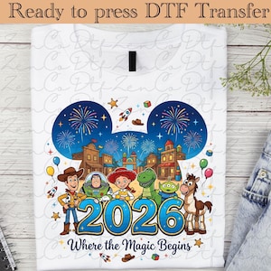 Peut inclure: T-shirt blanc avec un graphisme coloré. Le motif comprend des personnages de dessins animés, des feux d'artifice et un paysage urbain dans des oreilles de Mickey Mouse, avec le texte "2026 Where the Magic Begins."
