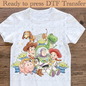 Peut inclure: T-shirt blanc avec un graphisme coloré des personnages de Toy Story, dont Woody, Buzz l'Éclair, Jessie, Rex, Zigzag, Bayonne et les Petits Hommes Verts. Le texte "Ready to press DTF Transfer" est en haut.