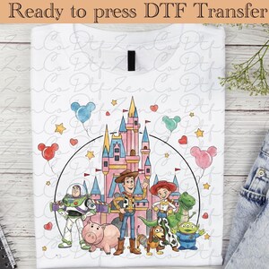 Peut inclure: T-shirt blanc avec un graphisme coloré des personnages de Toy Story, dont Woody, Buzz l'Éclair et Jessie, devant un château. Le motif comprend des ballons et des étoiles. Le texte "Ready to press DTF Transfer" est en haut.