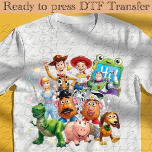 Peut inclure: Un t-shirt blanc avec une impression graphique colorée représentant divers personnages de jouets animés, dont un cow-boy, une cow-girl, un ranger de l'espace, un dinosaure vert, un cochon, un chien et deux figurines en forme de pomme de terre.
