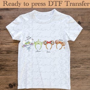 Peut inclure: T-shirt blanc avec des bandeaux de personnages de dessins animés : Buzz l'Éclair, Aliens, Jessie et Woody. Le t-shirt porte le texte "Ready to press DTF Transfer" en haut. Le t-shirt est sur une surface en bois.