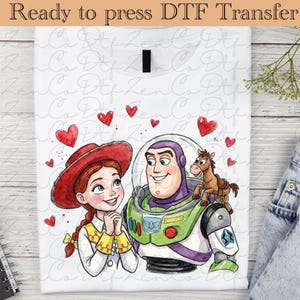 Peut inclure: T-shirt blanc avec une illustration à l'aquarelle de Jessie, Buzz l'Éclair et un petit cheval, entourés de cœurs rouges. Le texte "Ready to press DTF Transfer" est en haut.