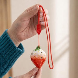 May include: A ceramic ocarina shaped like a strawberry, featuring red, white, and green colors. The instrument has several finger holes and is suspended by a red cord. The ocarina is held by a person.