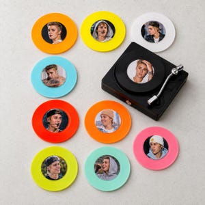 May include: A collection of colorful coasters featuring a pop star's image, alongside a miniature black turntable. The coasters are in various colors, including orange, yellow, blue, and pink. The turntable has a silver tone arm.