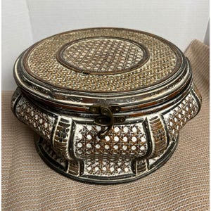 May include: A round, lidded storage box featuring a woven rattan pattern in shades of brown and white. The box has a dark brown wooden frame with decorative carved details and a central metal latch mechanism on the front to secure the lid.