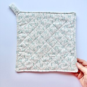 May include: A white quilted pot holder with a green floral pattern. The pot holder has a loop for hanging.