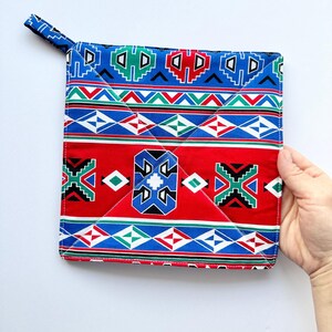 May include: A square fabric pot holder featuring a vibrant geometric pattern in red, blue, green, black, and white. The design includes horizontal bands of diamonds and stepped shapes, with a small fabric loop attached to the top corner for hanging.