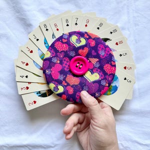 May include: A circular fabric card holder featuring a purple background with a multi-colored heart pattern and a bright pink button in the center. The holder is fanned out with a deck of standard playing cards showing various suits and numbers.