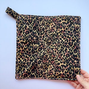 May include: A square, quilted pot holder with a loop for hanging. The fabric features a leopard print pattern in shades of black, brown, and gold. White stitching outlines the edges and creates a diamond pattern.
