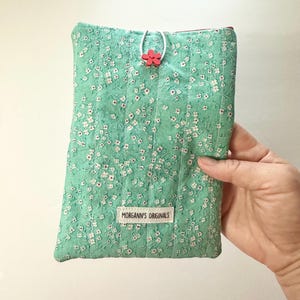 May include: A mint green quilted fabric case with a floral pattern of small white flowers. The case has a red flower closure and a label that reads "MORGANNS ORIGINALS".