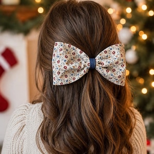 May include: A large hair bow with a floral pattern in cream, red, and blue is secured in the hair. The bow is attached to a dark blue fabric. The person is wearing a cream-colored sweater. The background is blurred with Christmas decorations.
