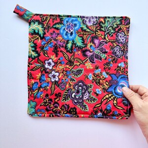 May include: A red quilted hot pad with a floral pattern in shades of blue, purple, and green. The hot pad has a loop handle for easy grabbing.